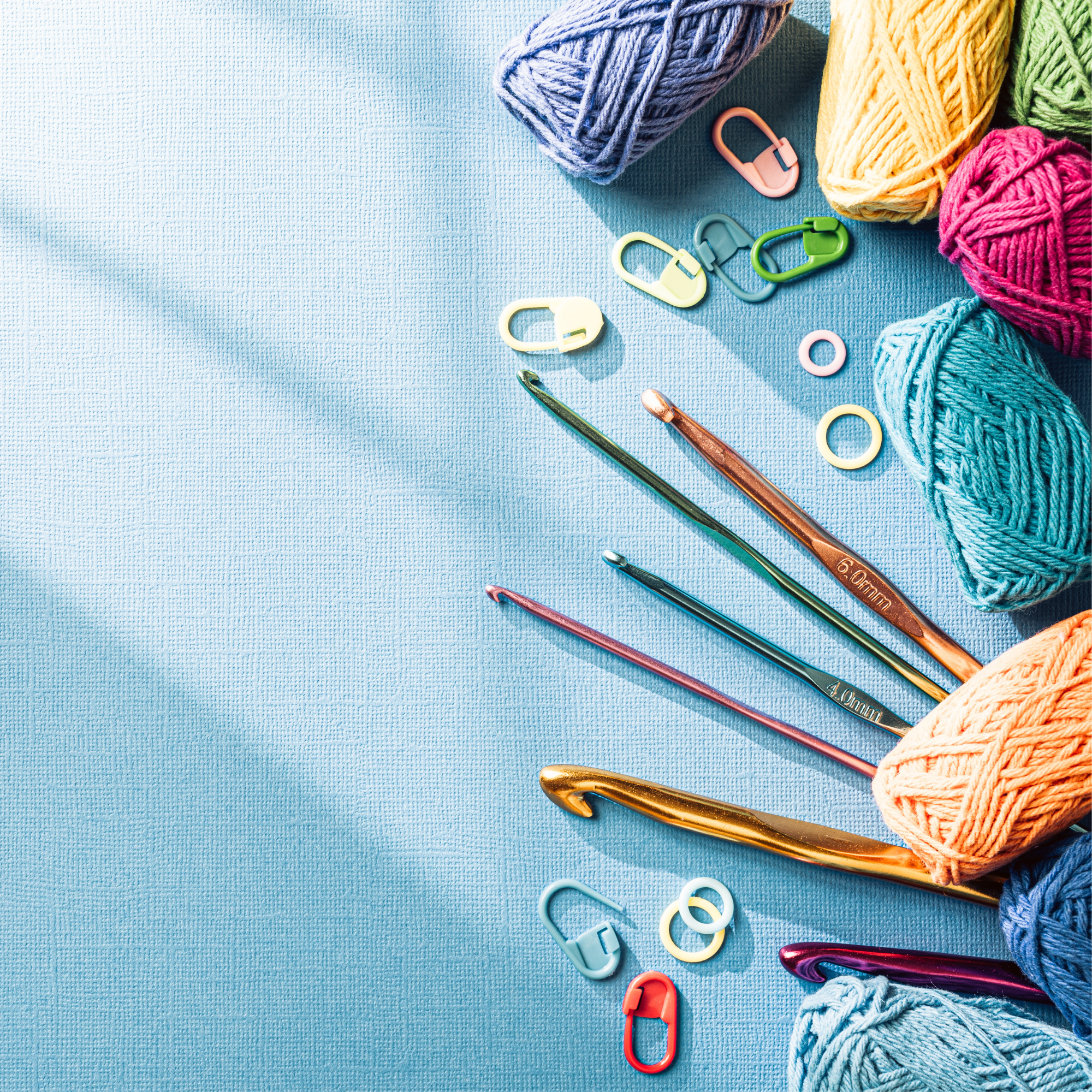 Blue background with crochet materials spread out, including crochet hooks, stitch markers, yarn, etc.