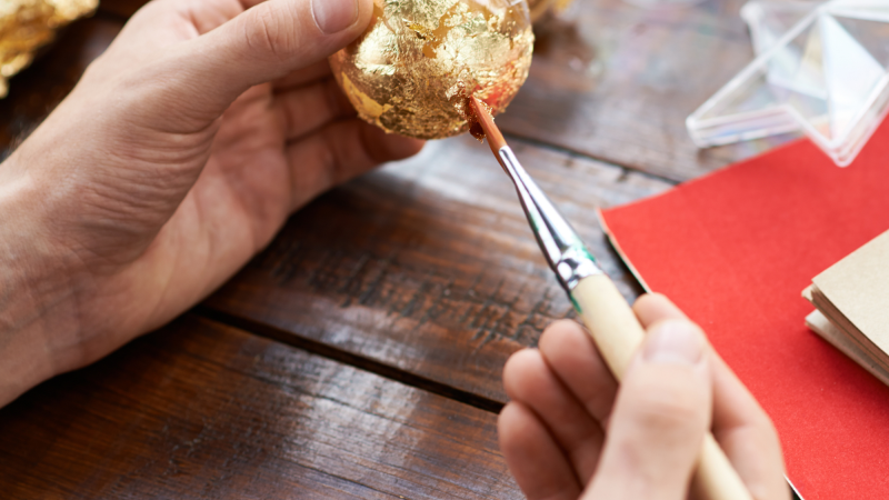 Photo showing hands applying gold foil to an ornament with other crafting materials in the background.